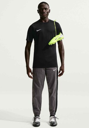 Mężczyzna w czarnej koszulce Nike i szarych spodniach Nike, trzymający na ramieniu jaskrawożółto-zieloną piłkarską korkociąg z sznurówkami.