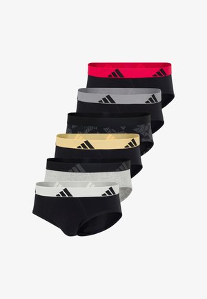 Athletischer dunkelhäutiger Mann steht barfuß da, trägt schwarze Badehose mit breitem gelbem Bund und schwarzem Adidas-Logo.