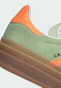 Sneaker Adidas con tomaia in suede verde e pelle arancione, suola traslucida color tan e logo impresso. Include dettagli con colletto imbottito.