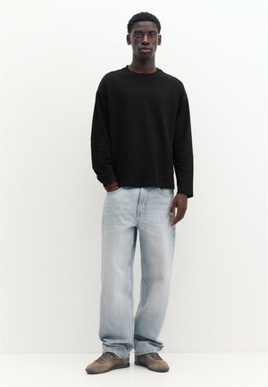 Ung mand stående mod hvid baggrund iført sort langærmet skjorte, lyseblå vidbenede jeans og brune sneakers.