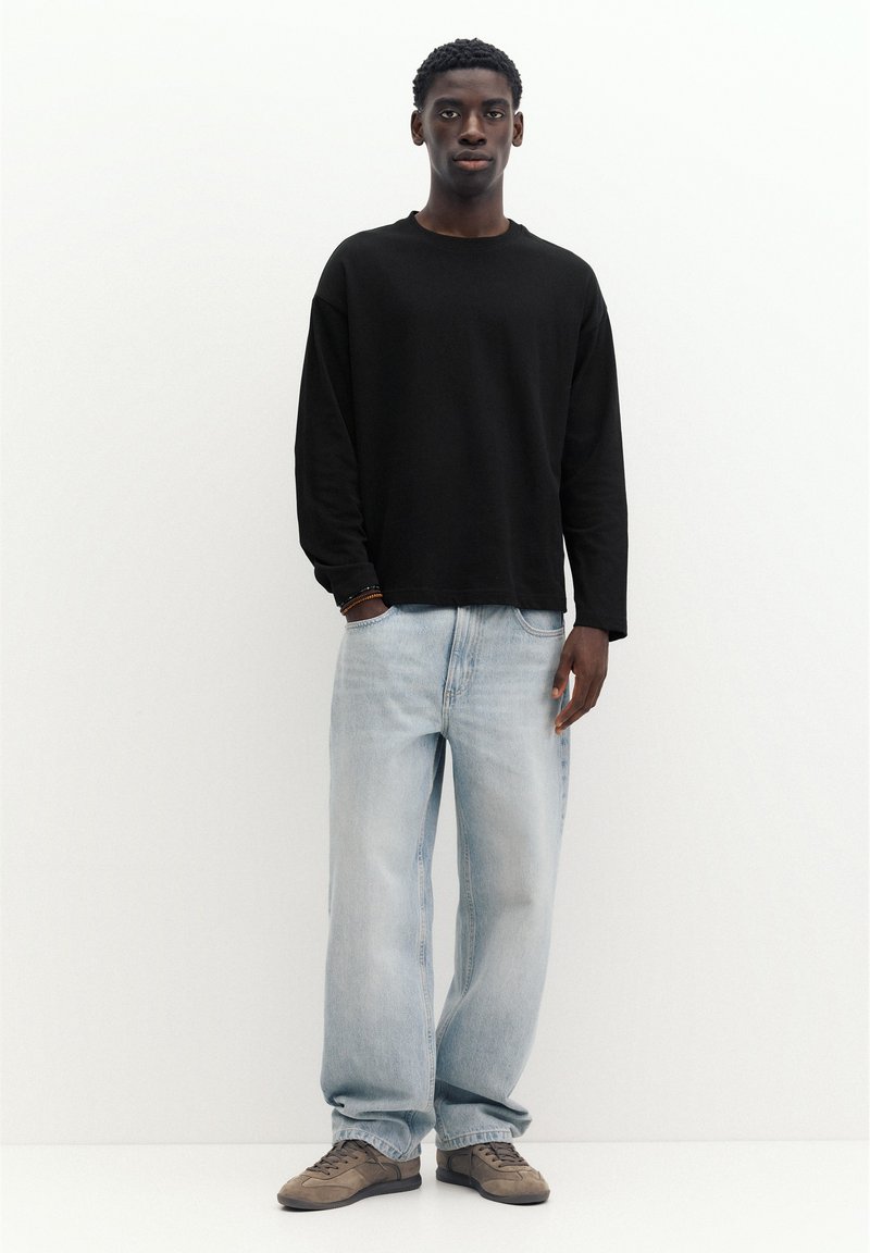 Jeune homme debout sur fond blanc, portant une chemise noire à manches longues, un jean large bleu clair et des baskets marron.