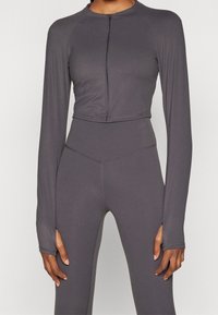 Šedá zkrácená sportovní bunda s dlouhými rukávy, zipovým uzávěrem a hladkou texturou, spárovaná s vysokým pasem odpovídajícími legínami.