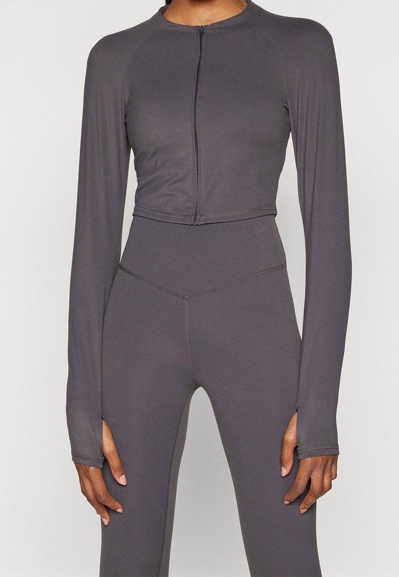 Šedá zkrácená sportovní bunda s dlouhými rukávy, zipovým uzávěrem a hladkou texturou, spárovaná s vysokým pasem odpovídajícími legínami.