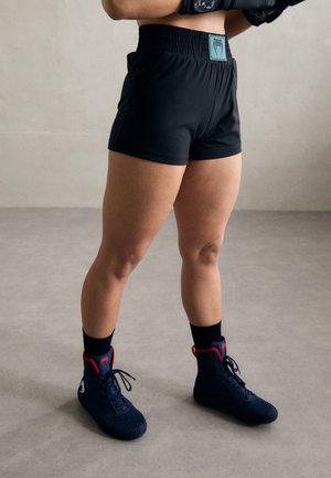 Person trägt schwarze Sportshorts, schwarze Socken und marineblaue Boxschuhe und steht auf einem grauen Boden vor einer einfarbigen Wand.