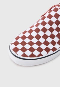 Gros plan sur l'avant d'une chaussure avec un motif damier marron et blanc et une semelle en caoutchouc blanche avec une bande noire sur fond clair.