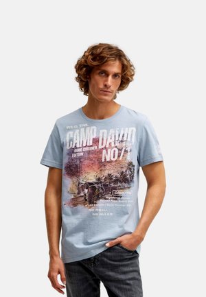 Junger Mann mit lockigem Haar, der ein hellblaues T-Shirt mit Wüsten- und Palmenaufdruck sowie Text trägt, steht mit einer Hand in der Tasche.