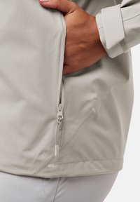 Lichtgrijze jack met een ritszak, gladde stof en verstelbare manchetten. Inclusief een trekkoord aan de zoom voor aanpassing.