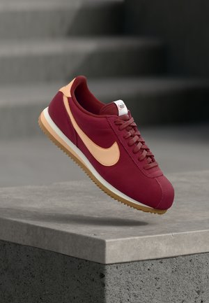 Vinrød ruskindsneaker med lyseorange Swoosh, teksturerede snørebånd og hvid sål; designet til letvægtskomfort og retroæstetik.