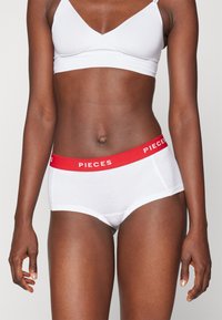 Hvite bomullstruser med en rød elastisk midjebånd som har "PIECES" i hvit tekst. Glatt tekstur og tettsittende design.