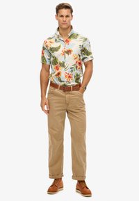 Camisa de manga corta con estampado floral en blanco, verde y rojo, combinada con pantalones beige y zapatos de cuero marrón. Cinturón marrón con detalle.