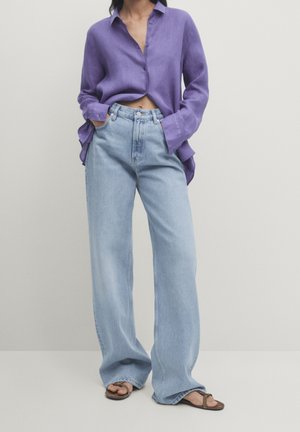 Persoon die losse lichtblauwe spijkerbroek draagt, een licht open paarse longsleeve en bruine sandalen tegen een effen lichte achtergrond.