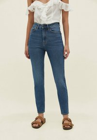 Blauwe spijkerbroek met hoge taille en een slanke pasvorm, voorzien van vijf zakken, een knoop- en ritssluiting, en bruine platte sandalen met twee gespen.