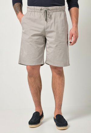 Mann trägt beige Shorts mit Kordelzug, schwarzes Langarmshirt und schwarze Slip-On-Schuhe mit geflochtenen Sohlen, steht mit einer Hand in der Tasche.