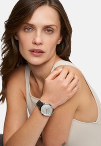 Silberne Armbanduhr mit rundem Ziffernblatt, schwarzem Metallarmband und schlichtem Design. Verfügt über ein lichtreflektierendes Ziffernblatt und dünne Stundenmarkierungen.