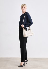 Beige leather handbag with a curved shape and gold hardware, paired with a navy textured long-sleeve top and black slim-fit pants.