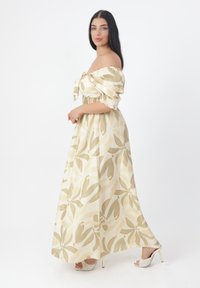 Robe maxi décolletée en beige avec un motif floral vert et jaune, taille élastique et tissu texturé ; portée avec des sandales à talons hauts.
