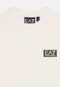 Balta kokvilnas T-krekls ar ovālu izgriezumu un mazu melnu logo uzģērbuma uzlīmi ar uzrakstu "EA7 EMPORIO ARMANI" kreisajā augšējā malā.
