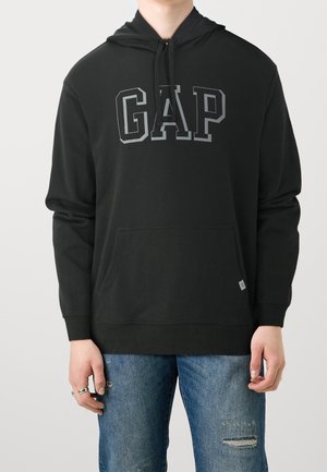 Persona que lleva una sudadera con capucha negra con el logo "GAP" en letras grises y vaqueros azules desgastados, de pie contra un fondo claro y liso.