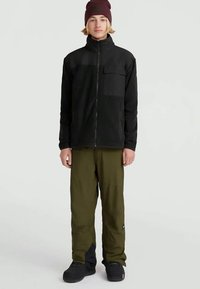 Schwarze Reißverschlussjacke mit strukturiertem Muster, olivgrüne Hose und schwarze Schuhe. Die Jacke verfügt über eine Brusttasche und ein tailliertes Design.