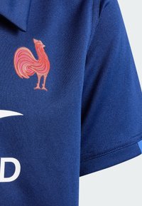 Modrá polo košeľa so zavedeným vyšitým logom ružového kohúta na ľavej strane hrudníka a bielym zakriveným dizajnom na prednej strane. Krátke rukávy.