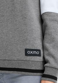 Šedá mikina s bílým rukávem a černým pruhem na spodním lemu. Obsahuje štítek "OXMO" na obdélníkovém černém podkladu. Měkká textura.