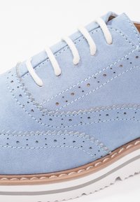 Chaussure en daim bleu clair avec des lacets blancs, présentant des détails perforés, des coutures en zigzag et une semelle extérieure texturée brunie avec des rayures blanches et grises.