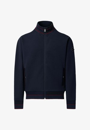 Chaqueta azul marino con cremallera y cuello alto, con franjas rojas en el cuello, los puños y el dobladillo, y bolsillos laterales con cierre de botón a presión.