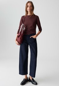 Donker bordeaux lange mouw top, wijde donkerdenim jeans en zwarte loafers. Een bordeaux tas met een gestreepte sjaal wordt in één hand vastgehouden.
