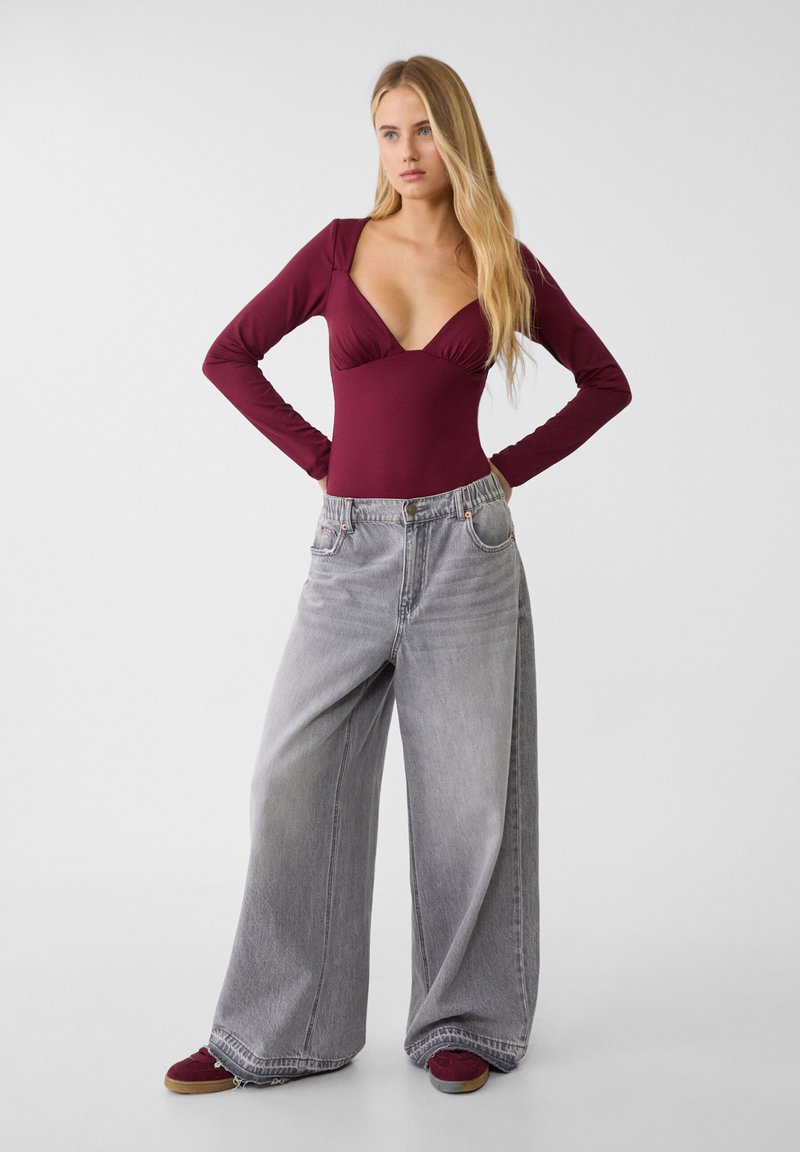 Body a maniche lunghe bordeaux con scollatura a V profonda e jeans grigi a gamba larga. L'outfit è completato da sneakers bordeaux con suola beige.