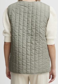 Gilet rembourré en vert clair avec des coutures verticales en forme de matelassage. Design sans manches avec un col et un dos arrondis. Accompagné d'un pull en tricot blanc.