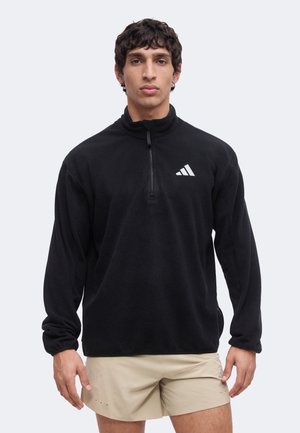 Mand iført sort halv lynlås fleece med Adidas-logo og beige shorts, stående mod en ensfarvet lys baggrund.