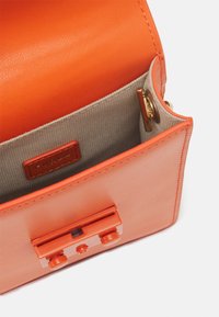 Pomarańczowa skórzana torebka otwarta, ukazująca beżowe wnętrze z tkaniny z małą skórzaną metką logo i złotym zapięciem.