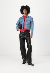 Veste en jean bleu clair avec des motifs blancs, associée à un haut rouge et un pantalon en cuir noir. Tenant un petit sac à main noir.