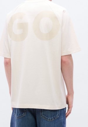Osoba nosząca beżowy t-shirt z krótkim rękawem z dużymi wyblakłymi literami "GO" na plecach i niebieskie dżinsy.