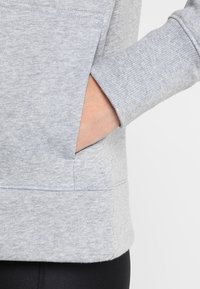 Hand gestoken in de voorzak van een lichtgrijze sweater met ribgebreide boorden, gedragen boven zwarte broek.