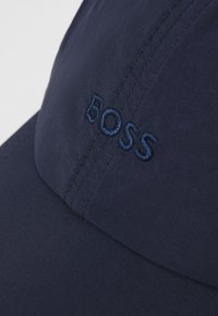 Tengerészkék sapka sima anyagból, amelyen egy kicsit világosabb tengerészkék hímzett "BOSS" logó található. Göndörített karima és látható varrási részletek jellemzik.