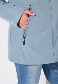 Ljusblå vattentät jacka med en dragkedjeficka, slät textur och rak passform. Detaljerad sömnad synlig längs fållen.