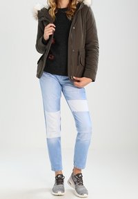 Oliwkowo zielona kurtka puchowa z kapturem obszytym futrem, czarny sweter, jasnoniebieskie jeansy z łatami oraz szare sportowe buty.