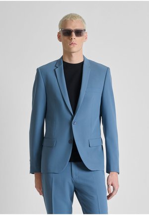 Giovane uomo che indossa un abito azzurro con una camicia nera e occhiali da sole scuri, in piedi contro uno sfondo chiaro e uniforme.