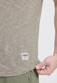 Kortærmet, meleret beige t-shirt lavet af blødt stof, med et hvidt mærke med sort tekst på den nederste side.