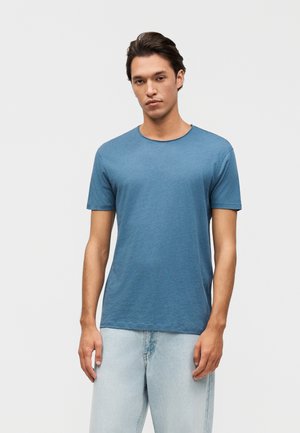 Mladý muž stojaci, oblečený v jednofarebnom modrom tričku s krátkym rukávom a svetlomodrých džínsoch, na jednoduchom svetlosivom pozadí.