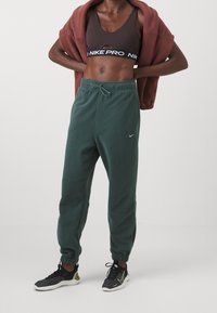Gröna fleece-joggers med elastiska ankle cuffs, snörning i midjan och subtil Nike-logotyp. Matchas med en mörkbrun sport-bh och svarta sneakers.