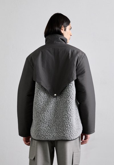 Grå fleecejacka med en slät, mörkgrå övre del. Har en flik bak, texturerad fleece och minimal hårdvara.