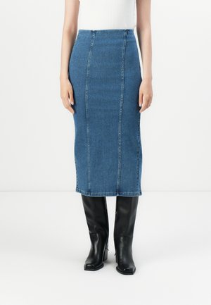 Woman wearing a fitted blue denim midi skirt with vertical seams and black knee-high heeled boots, standing against a white background.