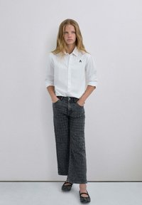 Camisa blanca de botones con un pequeño logo, combinada con vaqueros anchos negros de patrón y bailarinas negras. Material suave, diseño casual.