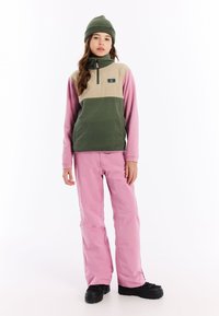 Fleecejack met groene, beige en roze kleurvlakken, half-rits kraag, gecombineerd met roze waterdichte broek en zwarte laarzen.