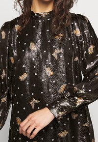 Femme portant un chemisier noir brillant à manches longues avec des pois blancs et des motifs de papillons orange, main reposant près de la taille.