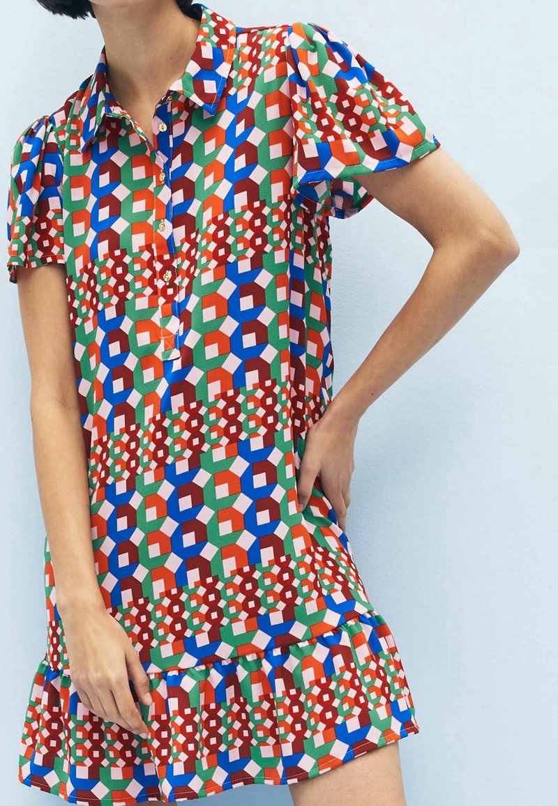 Femme portant une robe à manches courtes, à motif géométrique coloré, avec un col et une patte de boutonnage, se tenant devant un fond bleu clair.