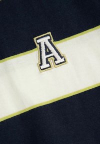 Granatowy dzianinowy materiał w kremowe i żółte poziome paski. Zawiera haftowaną literę "A" w białym kolorze z żółtą obwódką.