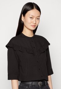 Blusa preta com um tecido texturizado, apresentando um colarinho franzido e botões na parte da frente, com mangas até ao cotovelo.